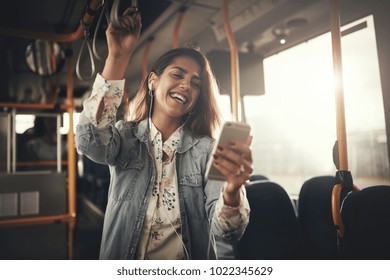 Young woman wearing earphones laughing at a text message on her cellphone while riding on a bus - Powered by Shutterstock