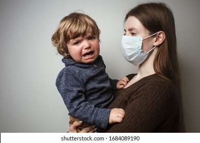 A Young Woman Wearing A Face Mask And Holding In Her Hands A Crying Toddler On A White Background. Protective Measures. Mother And Child In Quarantine. A Young Woman Holding A Baby Boy.