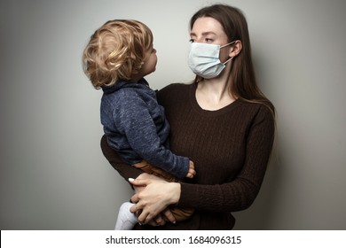 A Young Woman Wearing A Face Mask And Holding In Her Hands A Toddler On A White Background. Protection Against The COVID-19 Transmission. Mother And Child In Quarantine.