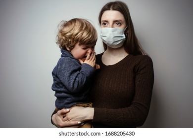 A Young Woman Wearing A Face Mask And Holding In Her Hands A Crying Toddler On A White Background. Protective Measures. Mother And Child In Quarantine. A Young Woman Holding A Baby Boy.