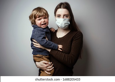 A Young Woman Wearing A Face Mask And Holding In Her Hands A Crying Toddler On A White Background. Protective Measures. Mother And Child In Quarantine. A Young Woman Holding A Baby Boy.