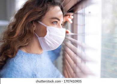 Young woman wearing a face mask and peeking out from blinds. Woman in quarantine looking through the window. Corona covid-19 virus protection. - Powered by Shutterstock