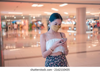 A young woman wearing a face mask inside a shopping mall checking her phone while walking around. - Shutterstock ID 2162110435