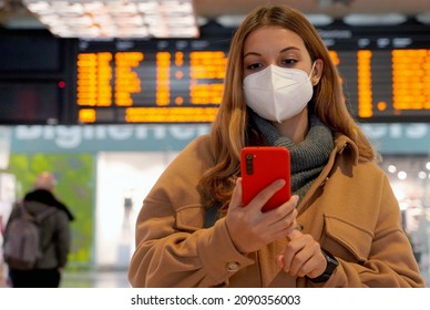 Young woman wearing FFP2 KN95 protective mask at train station. Female traveler checking her smartphone with behind timetables of departures arrivals.