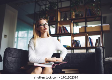 Young Woman Wearing Glasses And Casual Clothes Sitting On Sofa And Enjoying Interesting Books In Cozy Bookshop While Looking Away