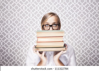 A young woman wearing glasses peers over a tall stack of books.