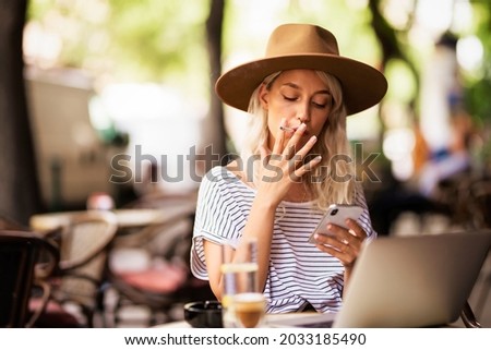 Young woman wearing hat while smoking a cigarette and text messaging. Fashionable woman sitting outdoors at the typical french cafe terrace.