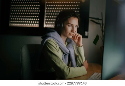 A young woman wearing headphones is focused working on her computer in a cozy office or at home. having a chat or watching a movie in a cozy home office at night  - Powered by Shutterstock