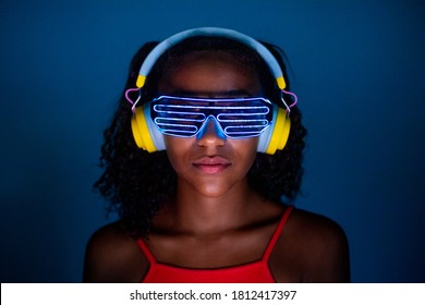 Young woman wearing headphones and futuristic led glasses on blue background - Isolated black woman wearing 3d smart glasses and headphones - virtual reality, future, technology concept 