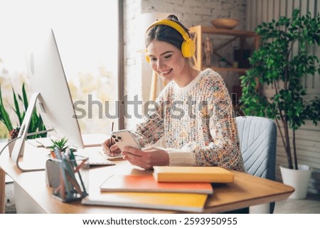 Young woman wearing headphones using smartphone and working at desk in a cozy home office interior during daylight