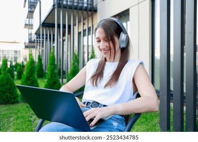 A young woman wearing headphones works on a laptop outdoors. She looks focused and relaxed in a modern urban setting. Perfect for remote work, digital nomad, and lifestyle productivity themes - Powered by Shutterstock