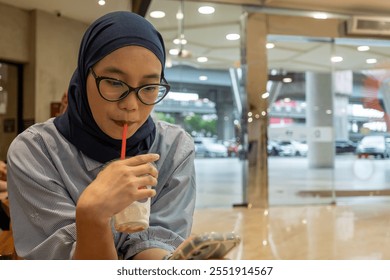 A young woman wearing a hijab is enjoying a drink in a modern café while using her smartphone. Perfect for themes of lifestyle, technology, or daily activities. - Powered by Shutterstock