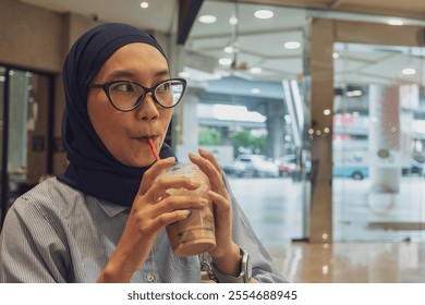 A young woman wearing a hijab is enjoying a drink in a modern café while using her smartphone. - Powered by Shutterstock