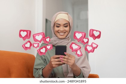 A young woman wearing a hijab is sitting on a couch and smiling as she uses her smartphone. Surrounding her are several pink like icons, signifying positive engagement on social media. - Powered by Shutterstock