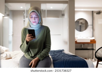 Young woman wearing a hijab sitting on a bed in a modern hotel room, engaging with her smartphone. The setting is comfortable and serene, highlighting technology use in everyday life.
 - Powered by Shutterstock