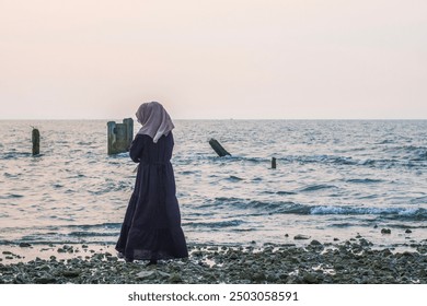 A young woman wearing a hijab is standing on the beach against the background of dim sunlight in the Pekalongan crematorium area. - Powered by Shutterstock