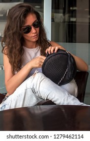 Young Woman In Wearing Jeans Looking For Something In Her Purse. Young Woman Looking Inside Her Bag. Trendy Girl Holding Small Nature Leather Bag In Hand. Stylish Accessories