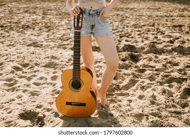 Young Woman Wearing Jeans Shorts, Standing Barefoot On Sand And Holding An Acoustic Guitar. 