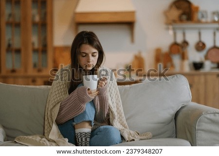 Young woman wearing a knitted sweater sitting at home on the sofa wrapped in a plaid drinking hot coffee or tea. Frozen woman will warm up in a cozy living room.