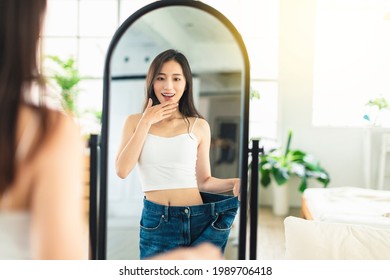 young woman wearing large-size jeans , looks at her figure in front of the mirror, very happy that she has succeeded in losing weight.