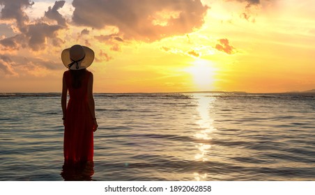 Young Woman Wearing Long Red Dress And Straw Hat Standing In Sea Water At The Beach Enjoying View Of Rising Sun In Early Summer Morning.