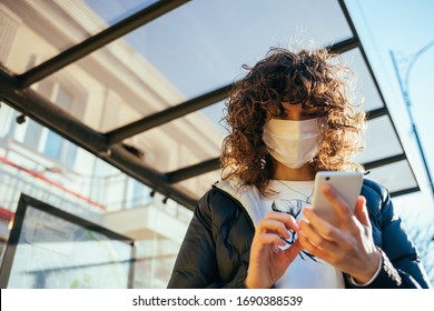 Young woman wearing medical face mask standing at public transport stop in European city and typing in her smart phone.