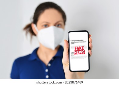 Young Woman Wearing Medical Mask And Holding A Smartphone With Fake News In Front. Propaganda, Misinformation. Horizontal Photo. Online Fake News On A Mobile Phone. 