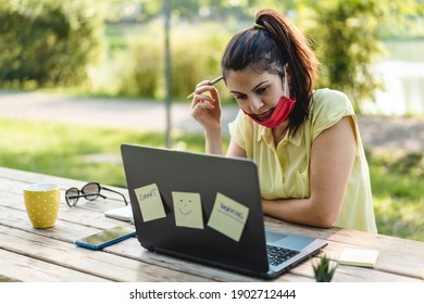 Young woman wearing open protective face mask using laptop outdoors - Pensive female solopreneur working in alternative workspace at park - Remote working, new normal concept after covid-19 pandemic