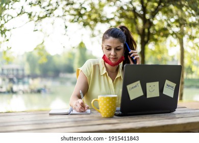 Young woman wearing open protective face mask using laptop outdoors - Female entrepreneur taking notes while calling with cell phone at park - Remote work, new normal concept after covid 19 pandemic