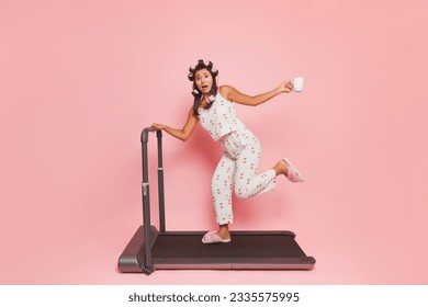 Young woman wearing pajamas in morning rush, running on treadmill, drinking coffee, curling hair with curlers, lifestyle concept, copy space - Powered by Shutterstock