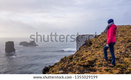 A young woman wearing a pink jacket, standing at the edge of a steep cliff. Some rock formations poking out of the sea. Moody atmosphere. Pure happiness and enjoyment derived from traveling