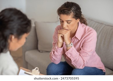 A young woman, wearing a pink shirt and jeans, sits on a beige couch, looking down with her hands clasped together in her lap. She appears to be in a counseling session - Powered by Shutterstock