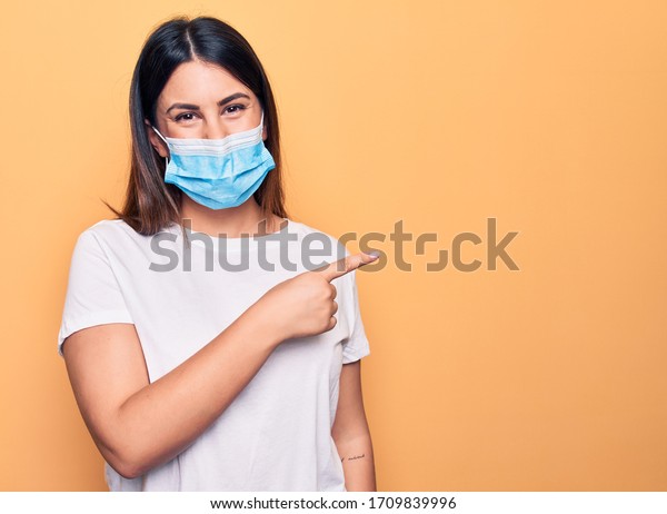 Young woman wearing protection mask for coronavirus\
disease over yellow background smiling cheerful pointing with hand\
and finger up to the side