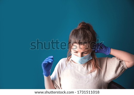 Young woman wearing protective gloves and face mask inside a home in quarantine looking bored and sad, for Covid-19 Coronavirus, with blue background portrait
