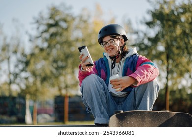 Young woman wearing protective helmet, using mobile phone and drinking takeaway coffee while sitting on skateboard at skatepark, enjoying break and sharing online content - Powered by Shutterstock