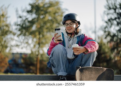 Young woman wearing protective helmet using her phone and drinking a coffee while sitting and resting on a ramp in a skatepark - Powered by Shutterstock