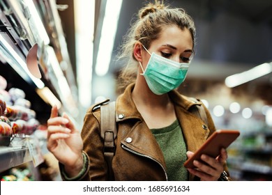 Young woman wearing protective mask on her face and reading shopping list on mobile phone in grocery store during virus pandemic.  - Shutterstock ID 1685691634
