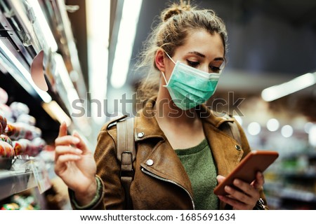 Young woman wearing protective mask on her face and reading shopping list on mobile phone in grocery store during virus pandemic. 