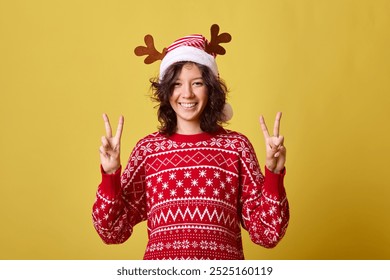 A young woman wearing a red sweater with a festive pattern and reindeer antlers headband is smiling and making peace signs with both hands. Isolated on a yellow background. - Powered by Shutterstock