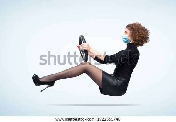 Young\
woman wearing a respiratory mask from coronavirus drives a car, on\
light blue background. Pandemic travel concept.\

