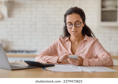 Young woman wearing round glasses, focused on her smartphone, seated at a wooden desk with laptop and documents, set against a brick-wall backdrop in contemporary kitchen setting.