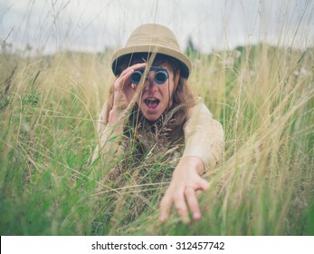 A young woman wearing a safari hat is hiding in the tall grass and is looking through binoculars