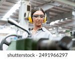 A young woman wearing safety glasses and ear protection is operating a lathe in a metalworking shop. looking at the workbench and serious.