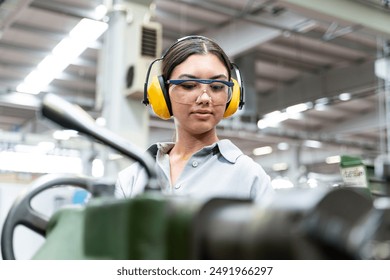 A young woman wearing safety glasses and ear protection is operating a lathe in a metalworking shop. looking at the workbench and serious. - Powered by Shutterstock