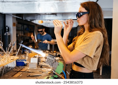 A young woman wearing safety glasses carefully blows glass with a torch in a studio setting, while another person works on a separate project nearby. - Powered by Shutterstock