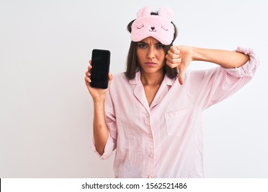 Young woman wearing sleep mask pajama holding smartphone over isolated white background with angry face, negative sign showing dislike with thumbs down, rejection concept