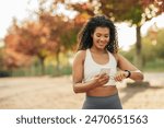 A young woman wearing sports attire is standing in a park during an autumn afternoon. She is looking at her fitness tracker while holding a smartphone, surrounded by colorful fall foliage.