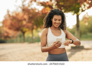 A young woman wearing sports attire is standing in a park during an autumn afternoon. She is looking at her fitness tracker while holding a smartphone, surrounded by colorful fall foliage. - Powered by Shutterstock