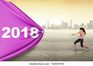 Young woman wearing sportswear while pulling numbers 2018 in a banner with city background - Powered by Shutterstock