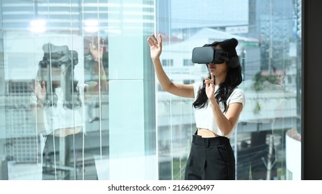 Young Woman Wearing Virtual Reality Headset And Interacts With Cyberspace, Standing Near Large Window With City Buildings View Outside